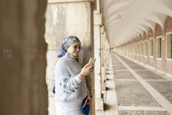 Young woman wearing headscarf using mobile phone while leaning on pillar