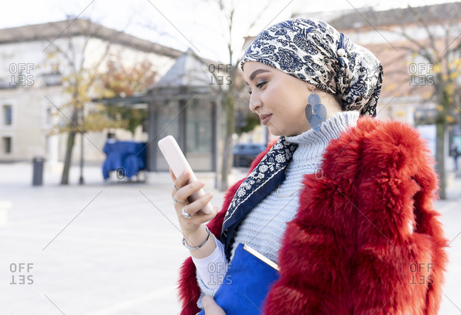 Smiling woman wearing headscarf and fur coat using mobile phone while standing in city