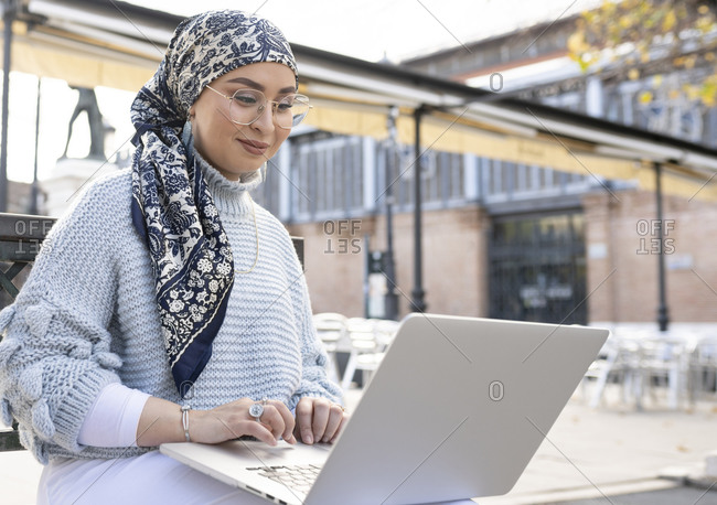 Woman wearing headscarf and eyeglasses working on laptop while sitting outdoors