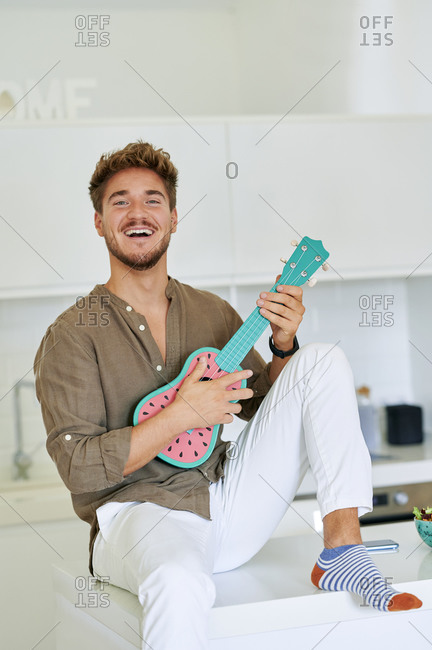 Cheerful young man playing ukulele while sitting on kitchen counter