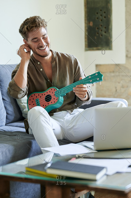 Smiling man learning ukulele through online tutorial on laptop at home