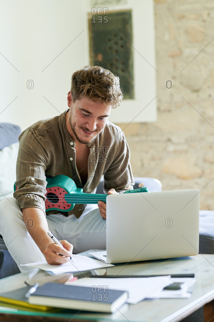 Young man writing on paper while learning ukulele online on laptop during in living room