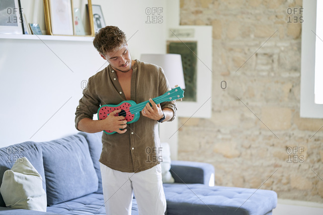 Male musician playing ukulele while standing in living room at home