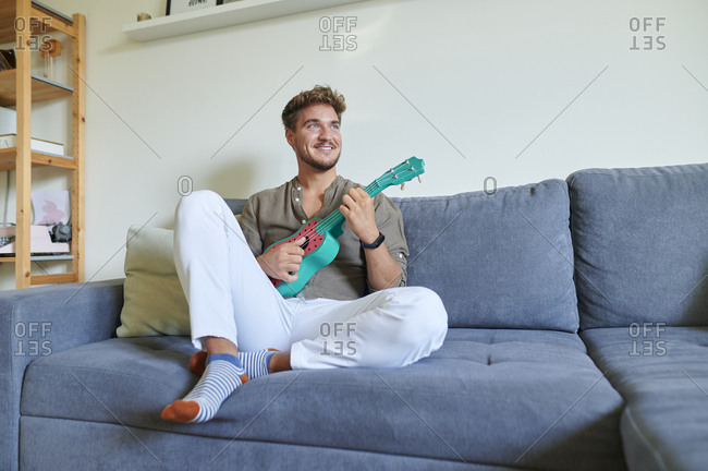 Smiling male musician playing ukulele while sitting on sofa at home