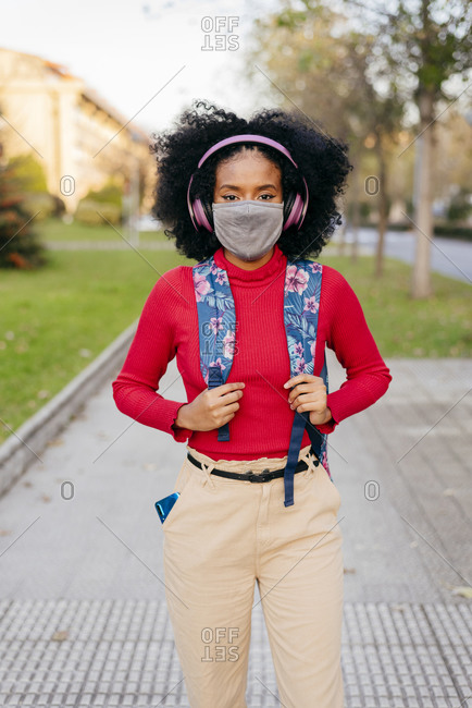 Woman wearing protective face mask and headphones carrying backpack while standing on footpath