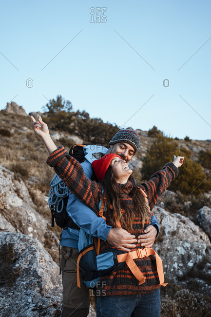 Happy boyfriend embracing girlfriend standing with arms outstretched on mountain during vacation