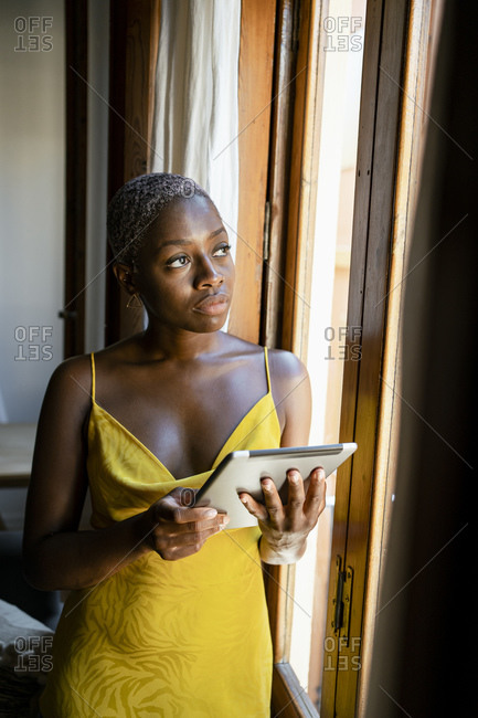 Thoughtful female hipster holding digital tablet while looking through window at home