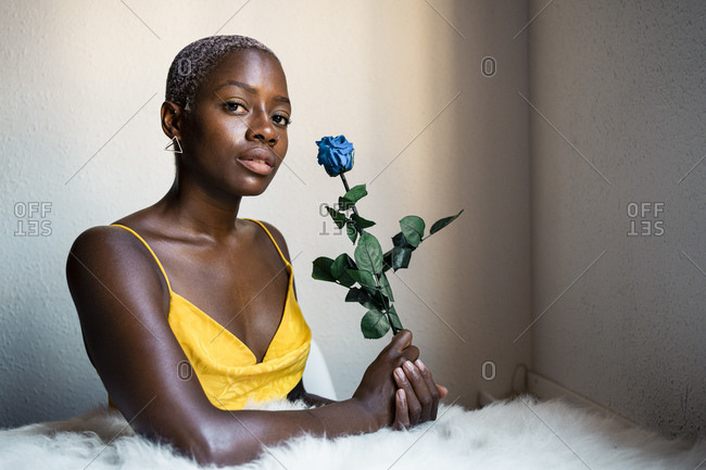 Confident female hipster sitting with blue rose against wall at home