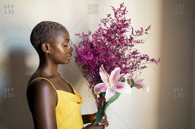 Woman in yellow dress smelling flowers by white wall at home