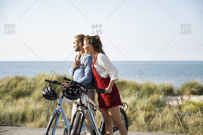 Girlfriend with arm around boyfriend admiring sea view while standing with bicycles at beach
