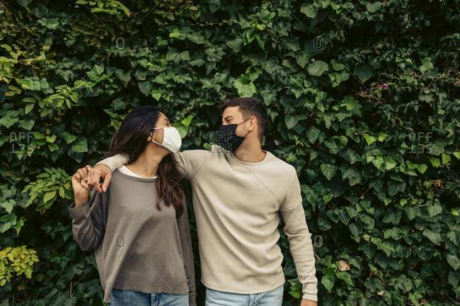 Young couple with arm around standing against leaves in park during pandemic