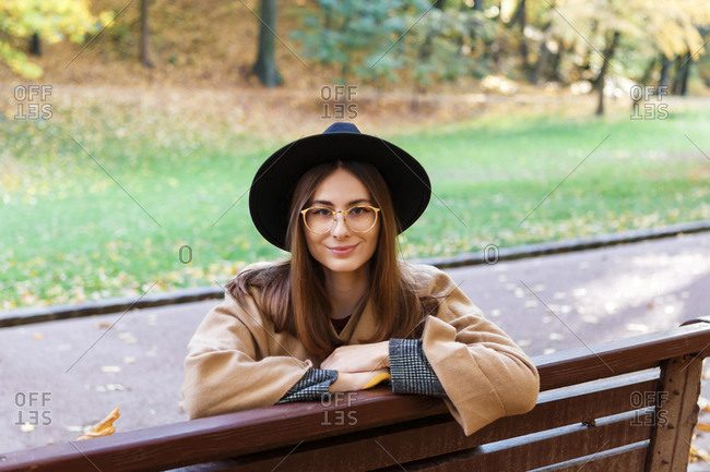 A young woman in a black hat and poncho is sitting on the bench in the park in the autumn season