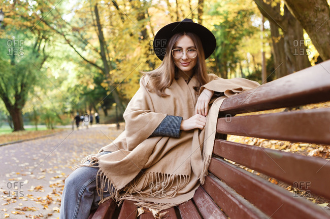 A young woman in a black hat and poncho is sitting on the bench in the park in the autumn season