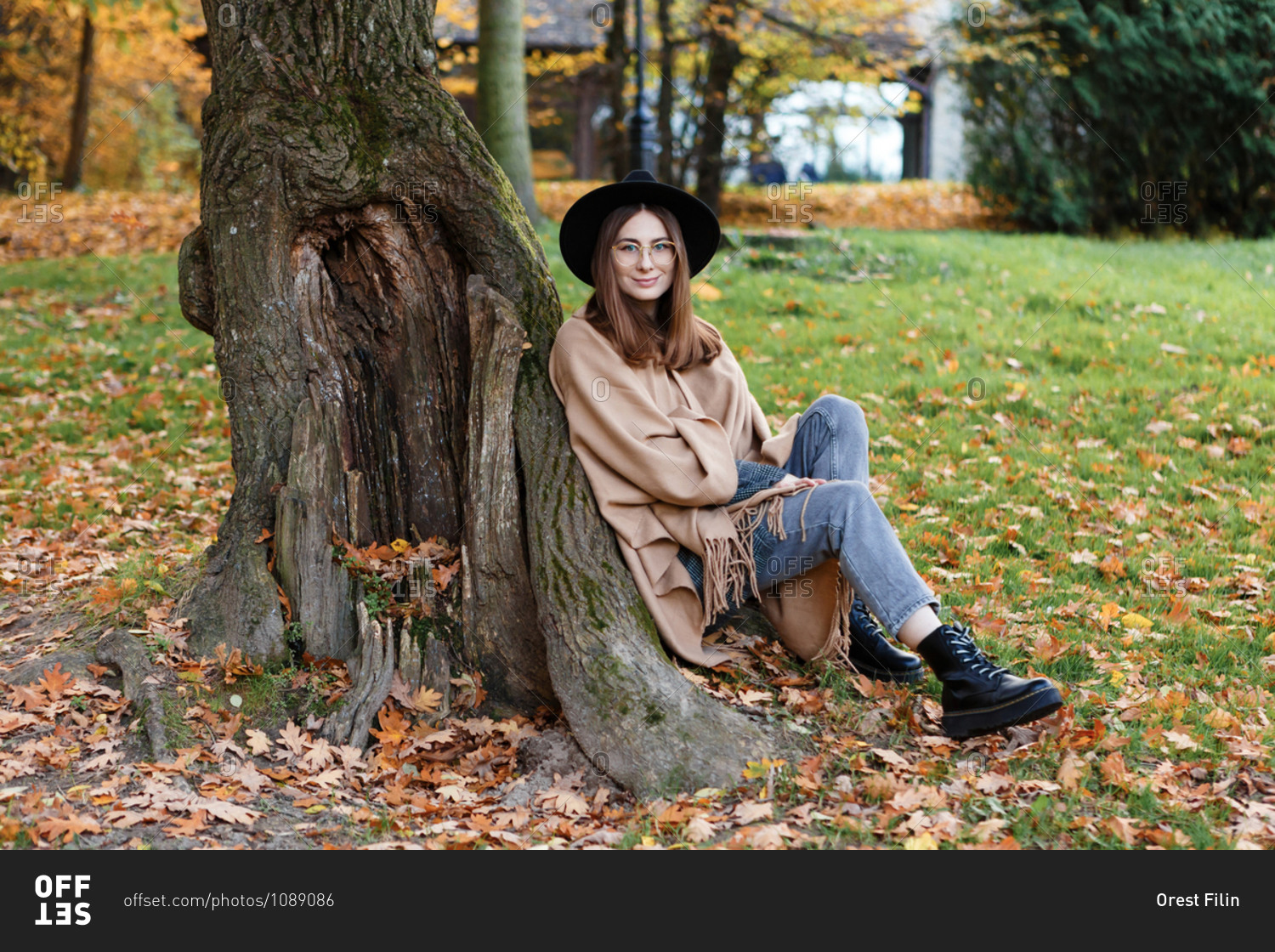 A young woman in a black hat and poncho is sitting under the tree on the grass in the park in the autumn season