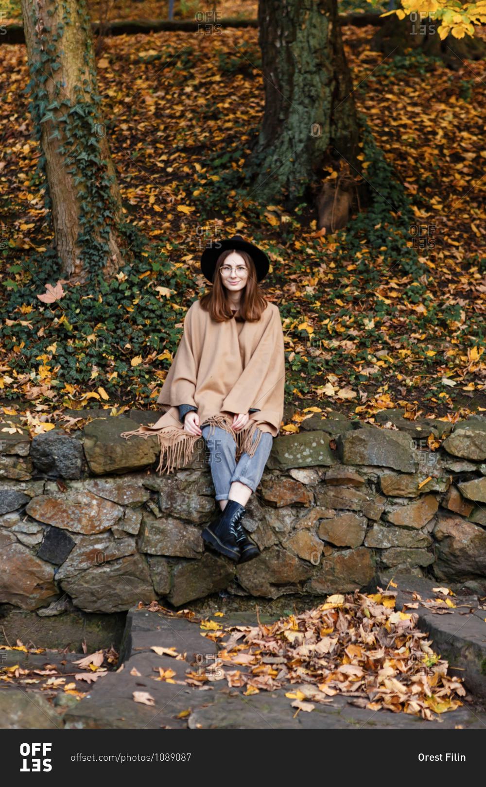 A young woman in a black hat and poncho is sitting on the small stone fence in the park in the autumn season