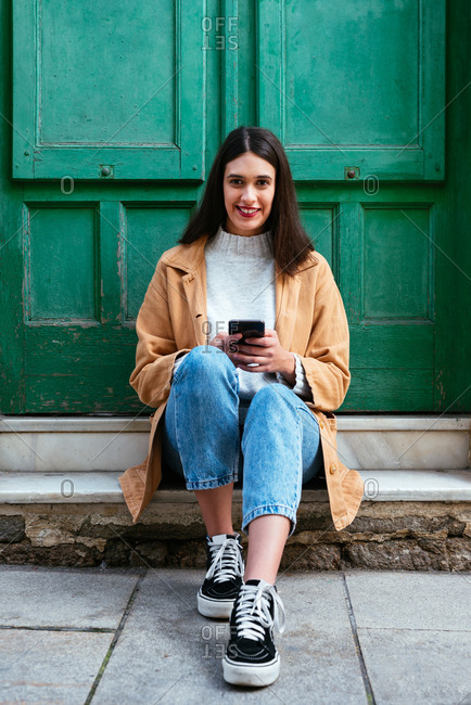 Portrait of young stylish woman sitting outdoors and using smartphone