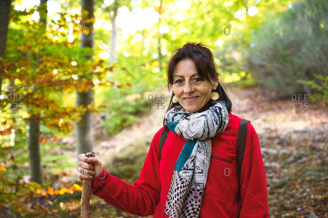 Smiling hiker woman holding a pole into forest while looking at camera