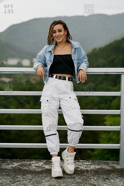 Young woman with blonde braided hair wearing a denim jacket and white jean resting on a bridge
