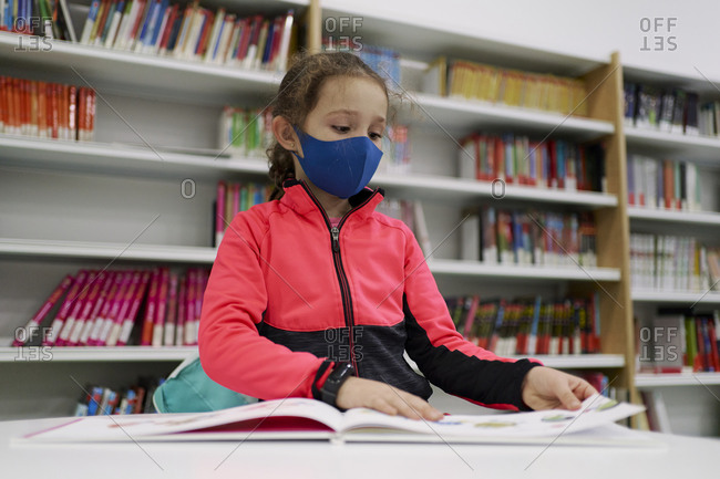 Little girl wearing a medical mask and reading a book in a libra