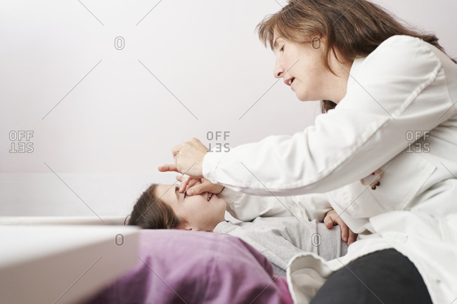 Female doctor smiling and giving eye drops to a little girl in her bed. Home doctor concept