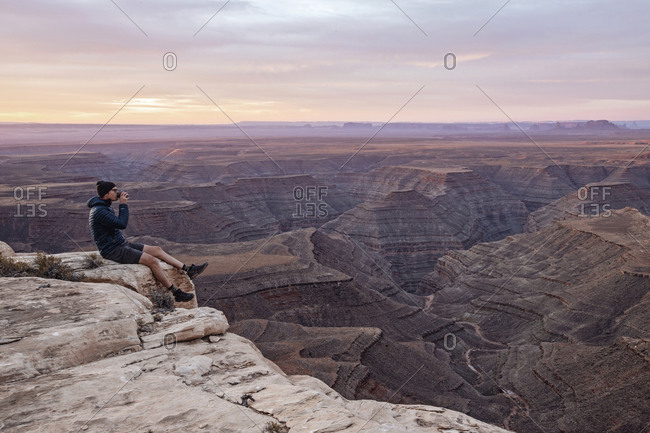 Man drinks coffee sitting on edge of cliff, Mexican Hat, Utah