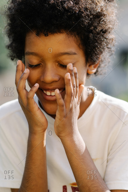 Portrait of happy little boy with curls covering eyes with hand, hiding closed eyes. Vertical shoot