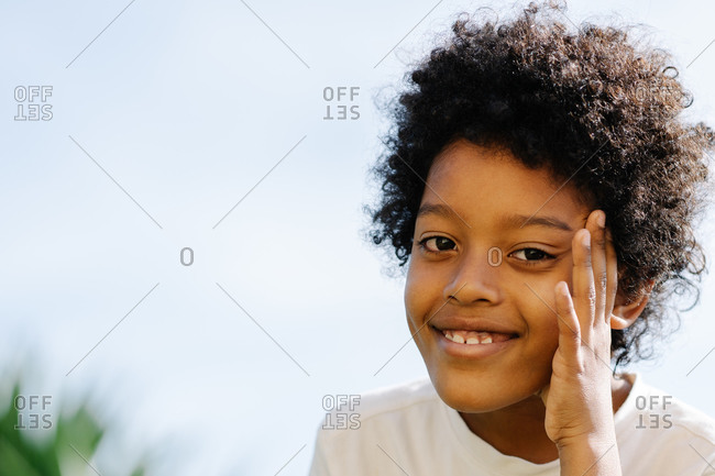 Afro nine year old boy keeping hand his face and looking at camera thoughtful happy facial expression