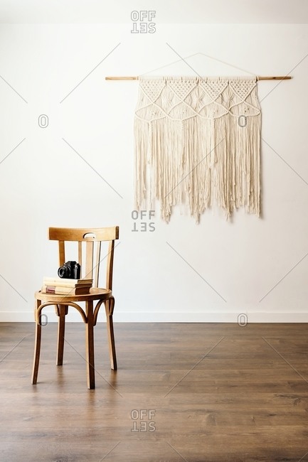 Minimalist interior of room with photo camera and books stacked on wooden stool placed against white wall with handmade boho hanging decoration