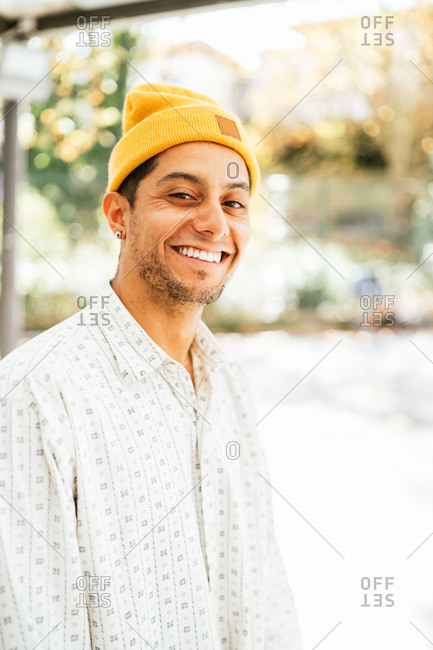 Positive young bearded hipster male in yellow hat and trendy informal shirt smiling and looking at camera while standing against blurred urban background