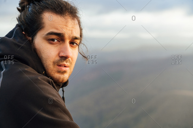 Lonely male explorer with backpack standing on top of hill and contemplating nature while traveling through mountainous terrain looking at camera