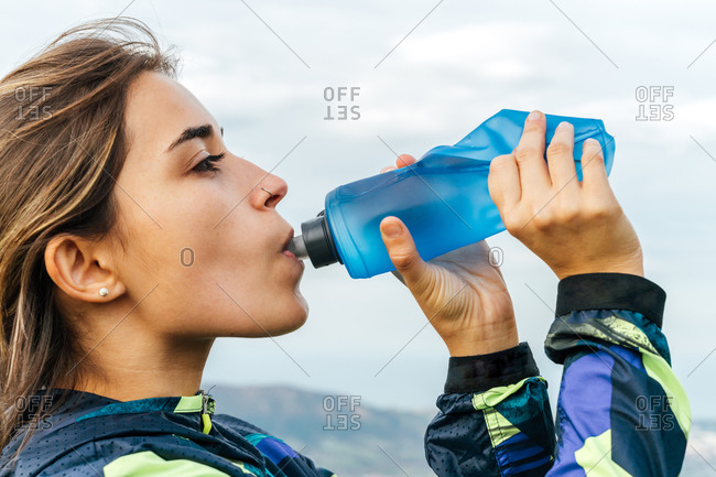 Side view of positive young ethnic female athlete in sportswear smiling and looking at camera while drinking water after workout in mountains