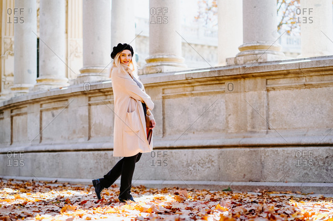 Side view of smiling female wearing coat and beret standing in autumn park near stone columns and looking away