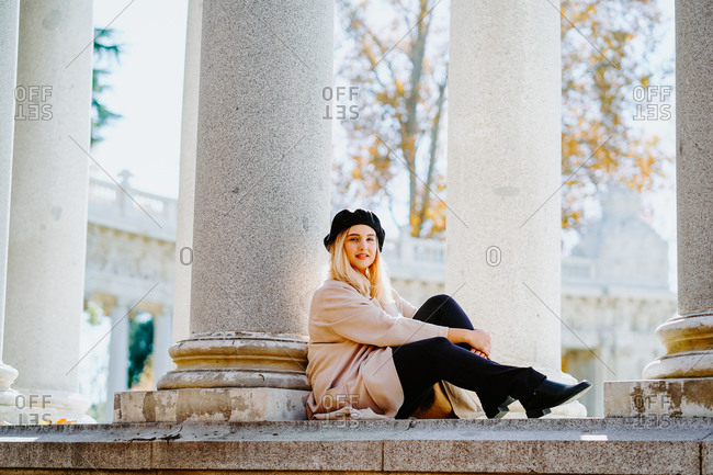 Side view of smiling female wearing coat and beret sitting near stone column in autumn park looking at camera