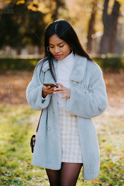 Ethnic female wearing autumn coat walking in park and messaging on social medial with friends via smartphone during stroll on sunny day