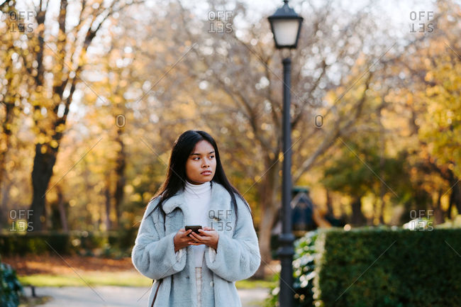 Ethnic female wearing autumn coat walking in park and messaging on social medial with friends via smartphone during stroll on sunny day