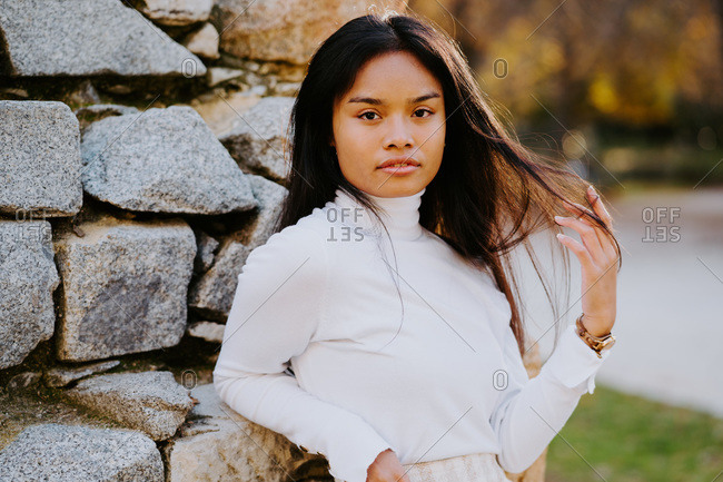 Carefree ethnic female in casual clothes leaning on stone building in park while touching hair and looking at camera