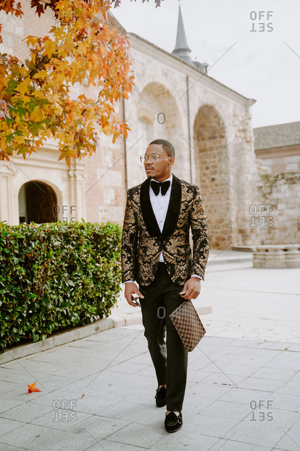 Elegant African American male wearing expensive tuxedo walking along street and looking away