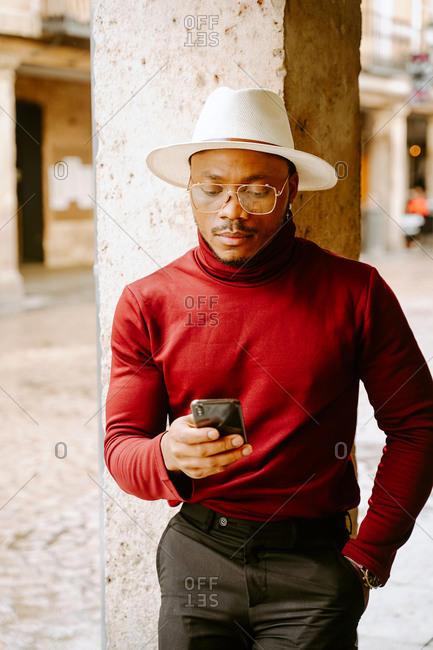 Determined African American male wearing elegant hat and clothes standing in street and browsing on smartphone while looking away