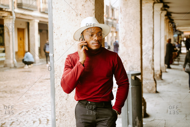 Determined African American male wearing elegant hat and clothes standing in street and speaking on smartphone while looking away