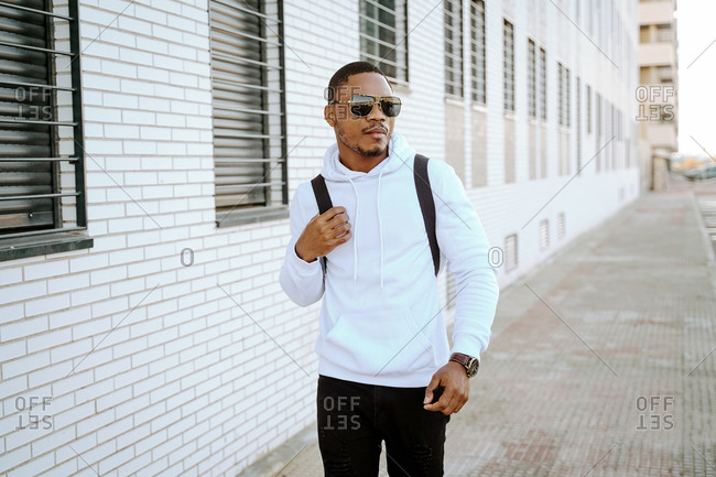 Cool African American male in stylish casual outfit walking on street and looking away