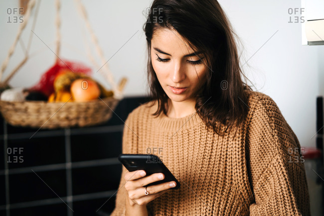 Relaxed female sitting with cup of coffee on counter in kitchen and browsing mobile phone while enjoying weekend at home