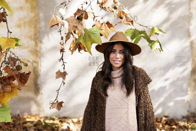 Fashionable young smiling ethnic female with long dark hair in stylish warm winter outfit and hat standing in autumn park near aged house with shabby walls looking at camera