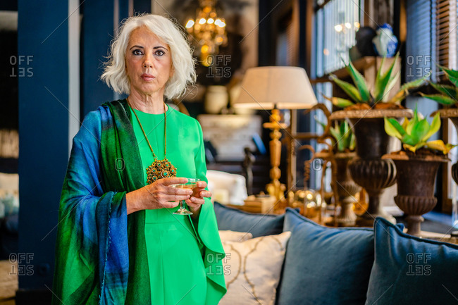 Mature female wearing green elegant dress standing with glass of alcohol drink in luxury house and looking at camera