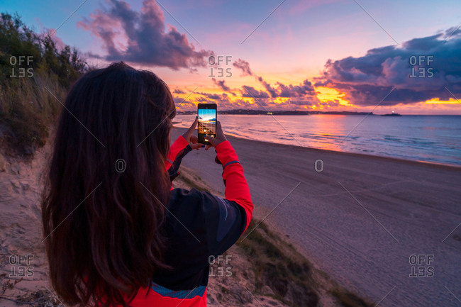 Back view of unrecognizable female traveler with long dark hair in casual clothes taking picture of amazing sunset over sea with sandy beach on smartphone during holidays in Cantabria