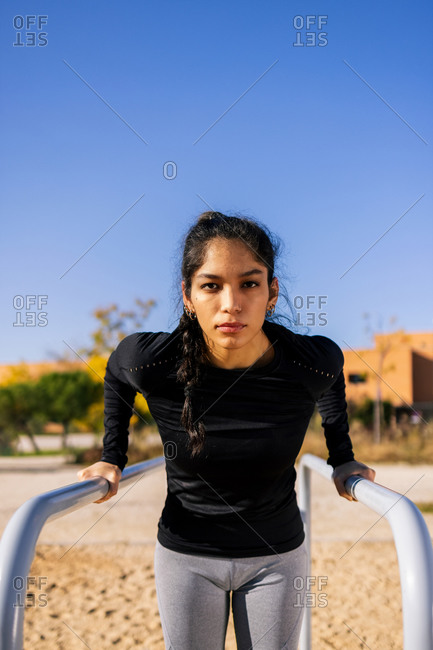 Confident slim ethnic female doing triceps exercise on parallel bars during calisthenics workout and looking at camera
