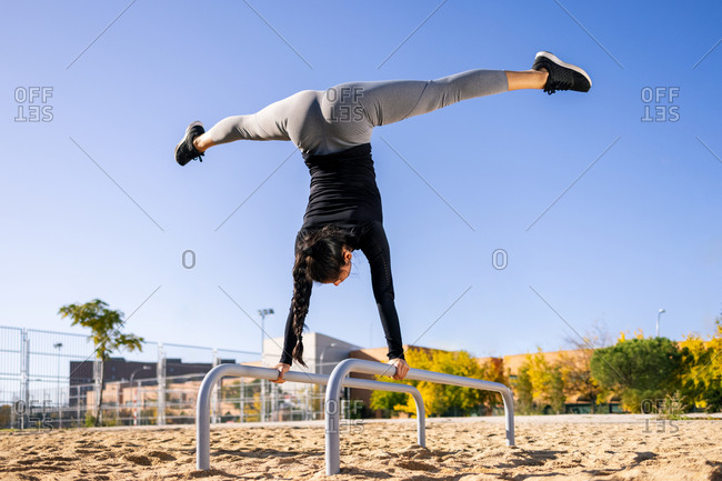 Back view of fit female athlete performing handstand with splits on parallel bars while doing exercises during calisthenics training
