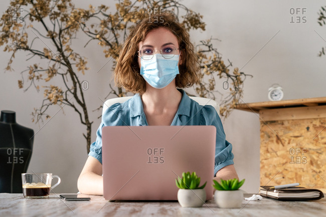 Concentrated young female entrepreneur in casual clothes and medical mask typing on laptop while working on project during quarantine. Looking at camera