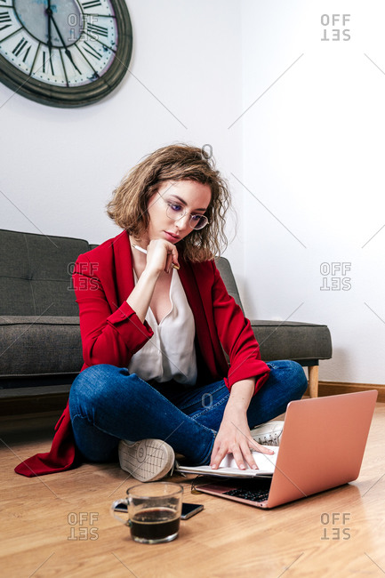 Full body of thoughtful young female student in casual outfit and eyeglasses sitting on floor near sofa and reading notes while doing homework assignment using laptop in modern apartment