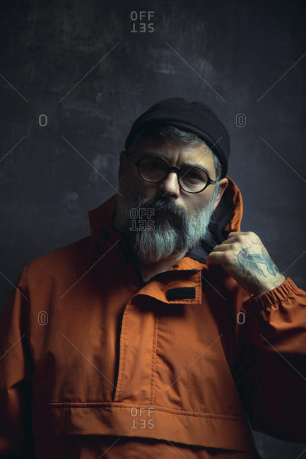 Pensive stylish tattooed adult ethnic male with gray beard in trendy hooded jacket and eyeglasses looking away against black background