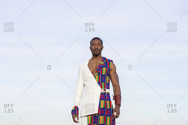 Determined African American male model in traditional costume standing on background of blue cloudy sky and looking at camera